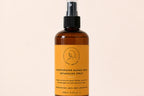 An amber glass spray bottle of Love Me Beauty Moisturising Mango Seed Detangling Spray with a black nozzle and a bright orange label, set against a solid light beige background.